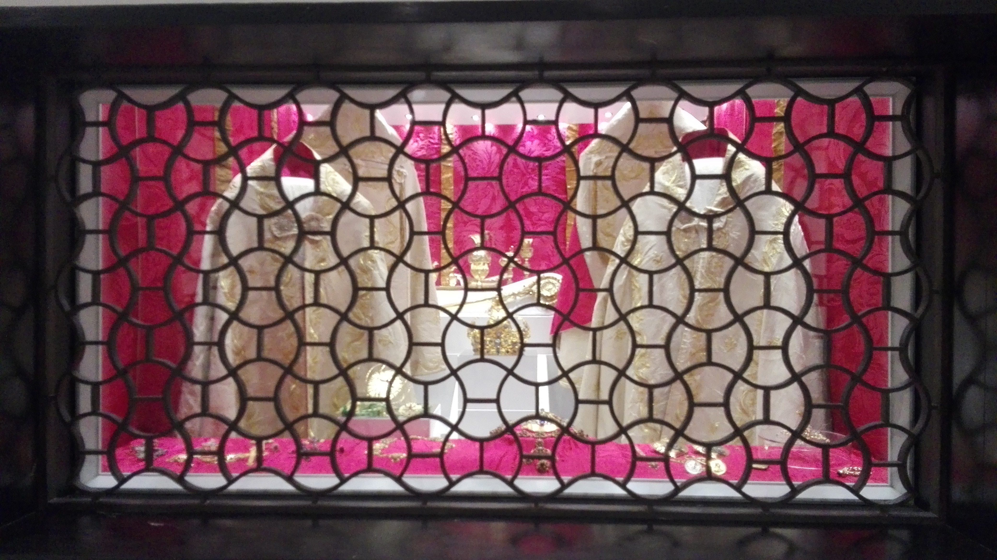 Esposizione di preziosi paramenti sacri vista da una grata decorativa.